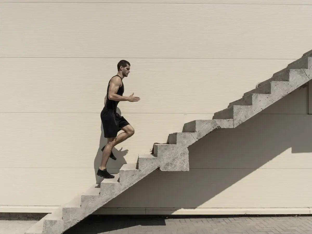 Mężczyzna w czarnym stroju sportowym wspina się po betonowych schodach na tle jasnej ściany.