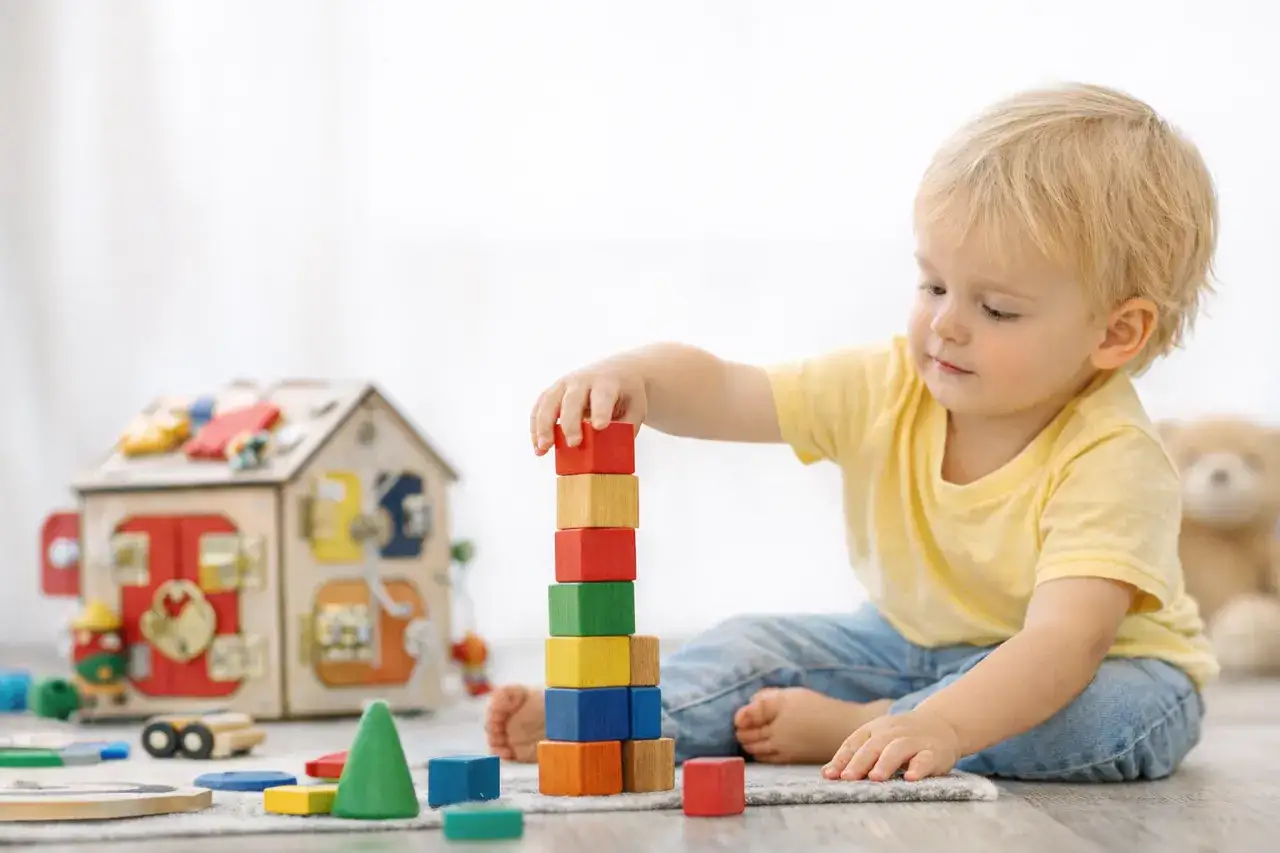 Chłopiec buduje wieżę z kolorowych klocków obok drewnianego domku.