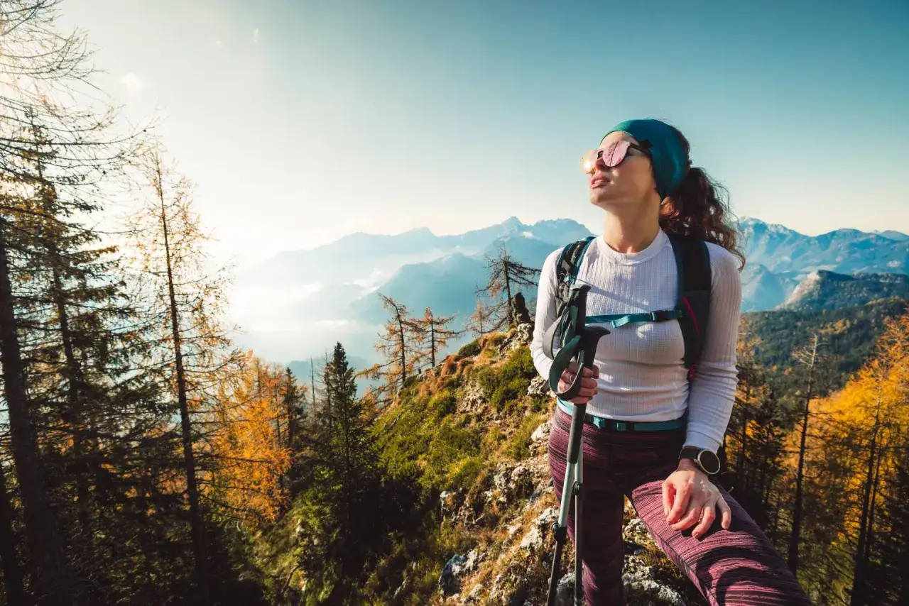 Kobieta w okularach przeciwsłonecznych i sportowym stroju, z kijkami trekkingowymi, podziwia jesienny krajobraz górski. Idealny przykład, jak się ubrać w góry.