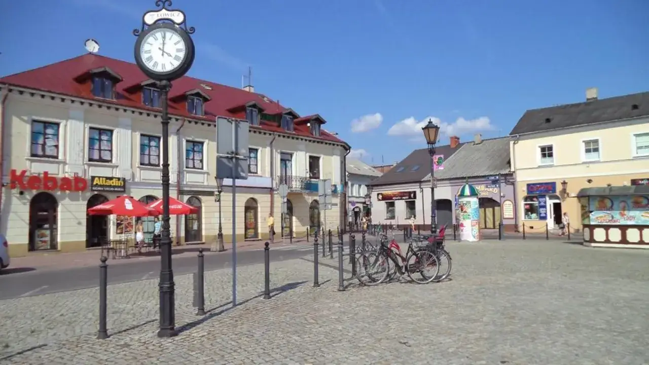 Słoneczny stary rynek łowicz z brukowanym placem, budynkami z czerwonymi dachami i zegarem na słupie.