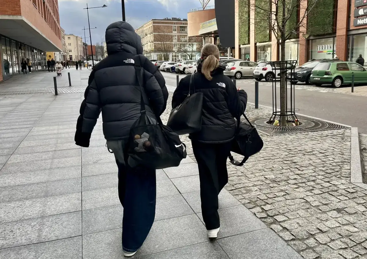 Dwie osoby w czarnych kurtkach The North Face idą chodnikiem, niosąc torby.