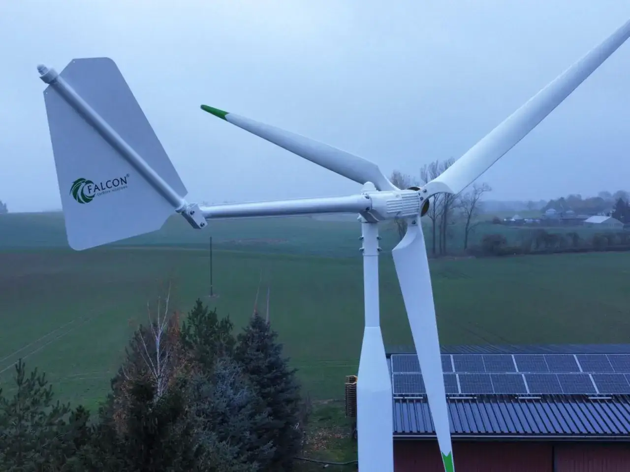 Biała turbina wiatrowa Falcon z zielonymi akcentami na tle pól i dachu z panelami słonecznymi.