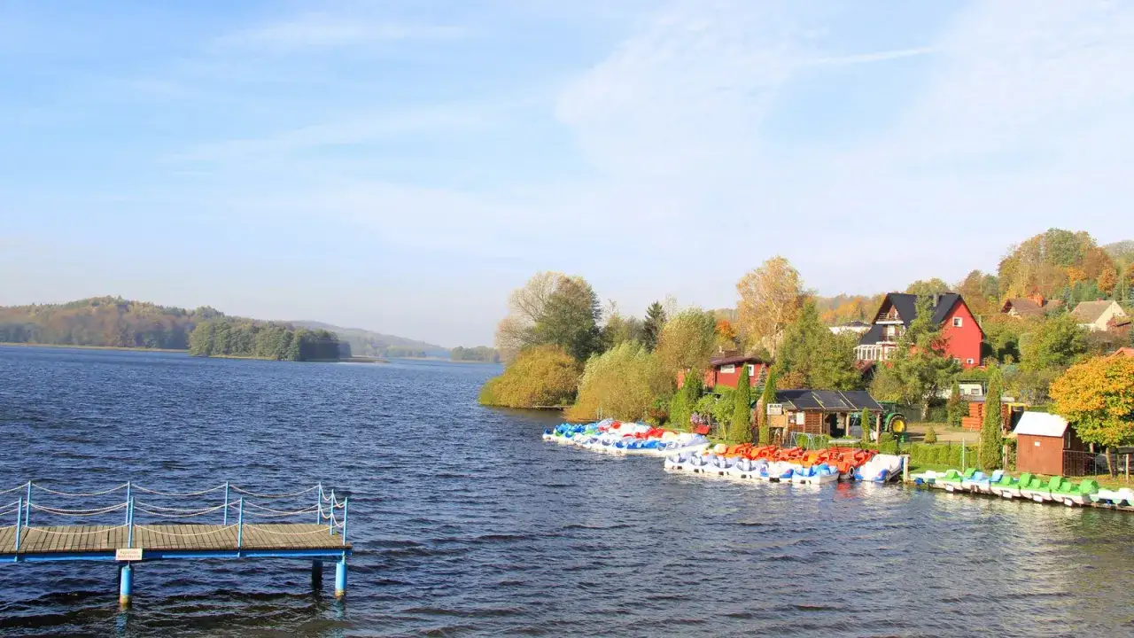 Widok na jezioro z pomostem i kolorowymi rowerami wodnymi przy brzegu, w tle domy i jesienne drzewa.