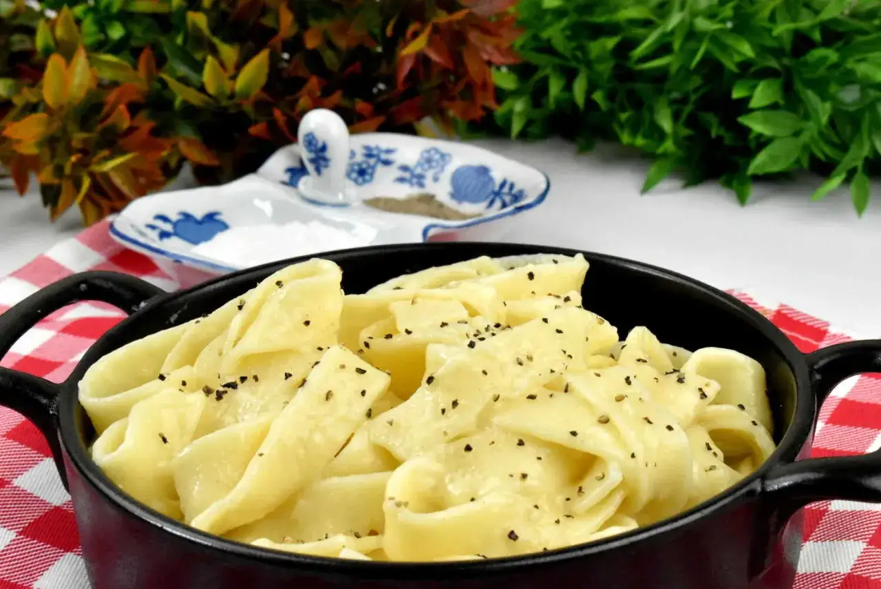 Pappardelle w kremowym sosie alfredo, posypane świeżo mielonym pieprzem, podane w czarnym naczyniu na czerwono-białej kratce.