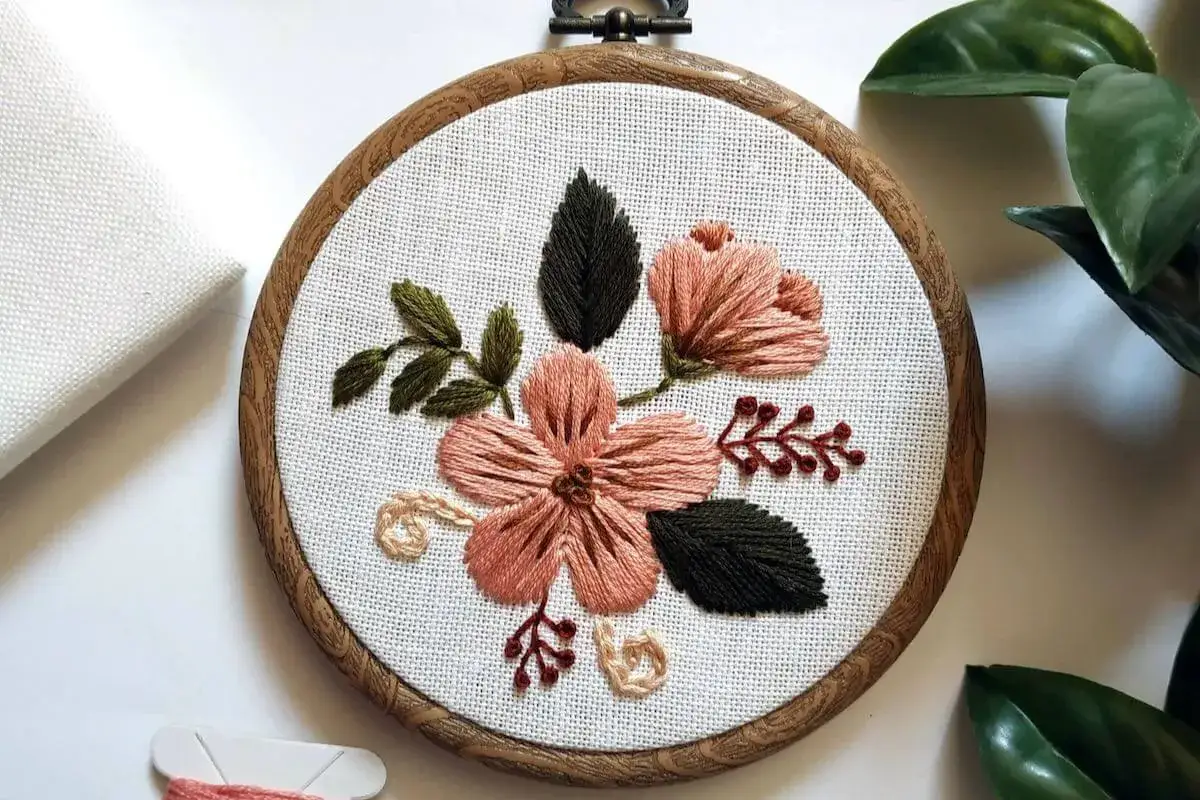 Kwiatowy haft na tamborku, ukazujący różnorodne techniki haftu. Różowe kwiaty i zielone liście tworzą delikatną kompozycję.