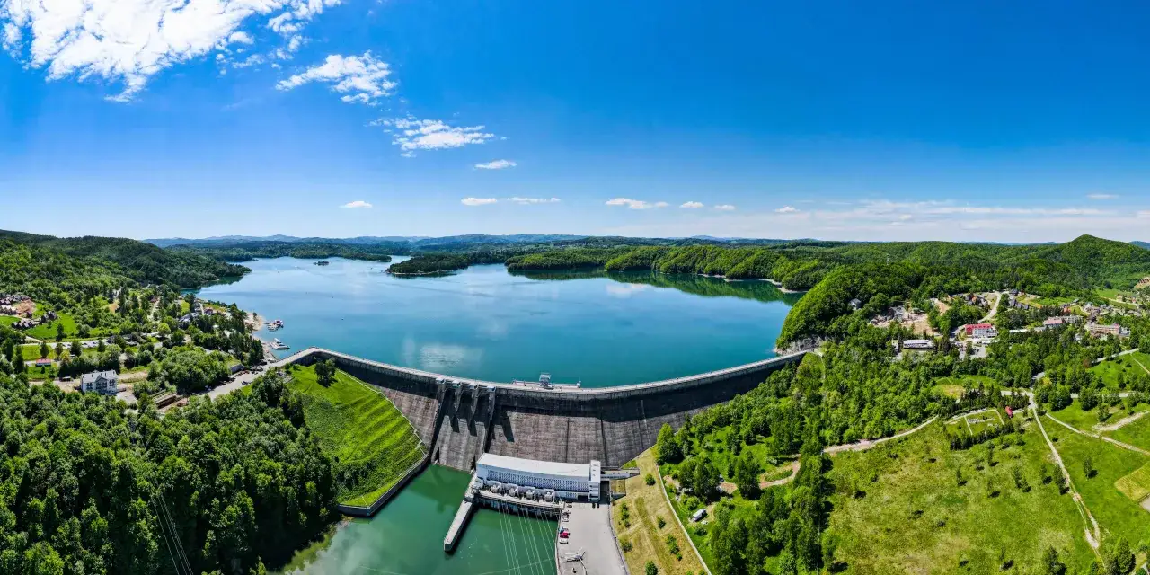 Zapora wodna z elektrownią, otoczona zielonymi wzgórzami i błękitnym jeziorem.