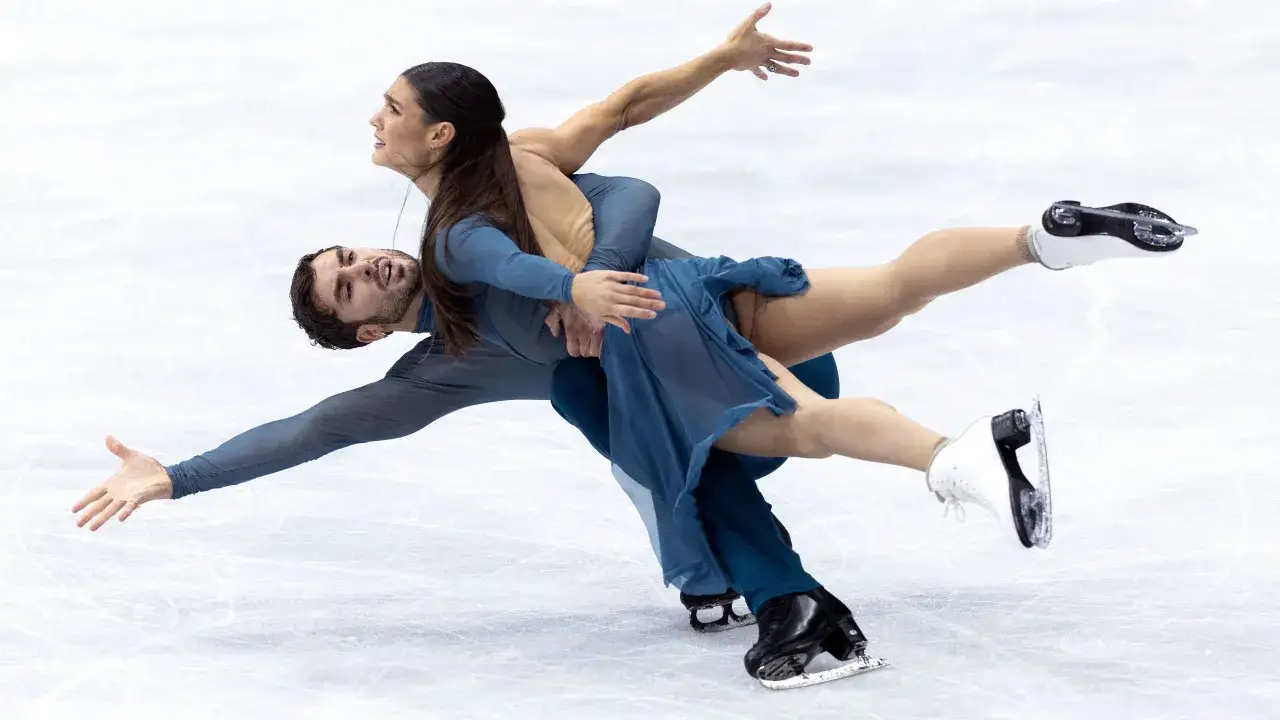 Para tańczy na lodzie w błękitnych strojach, wykonując skomplikowaną figurę. Ich ruchy to czysta jazda figurowa na lodzie.