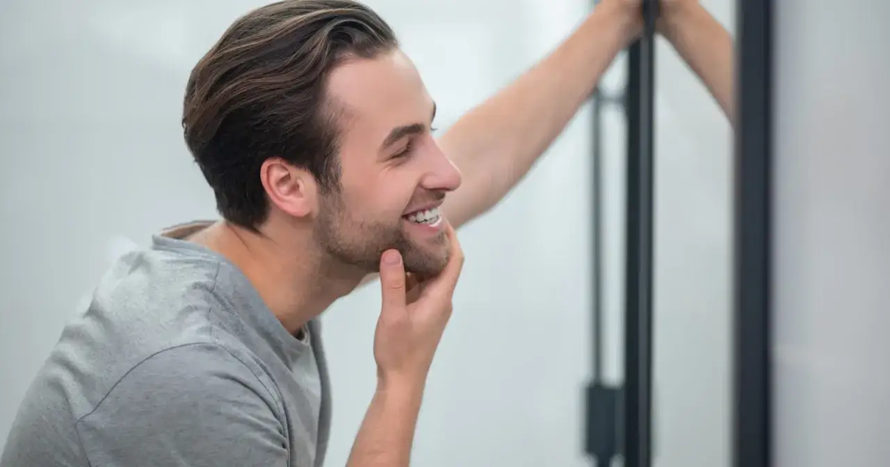 Mężczyzna w szarym t-shircie dotyka brody, uśmiechając się do lustra.