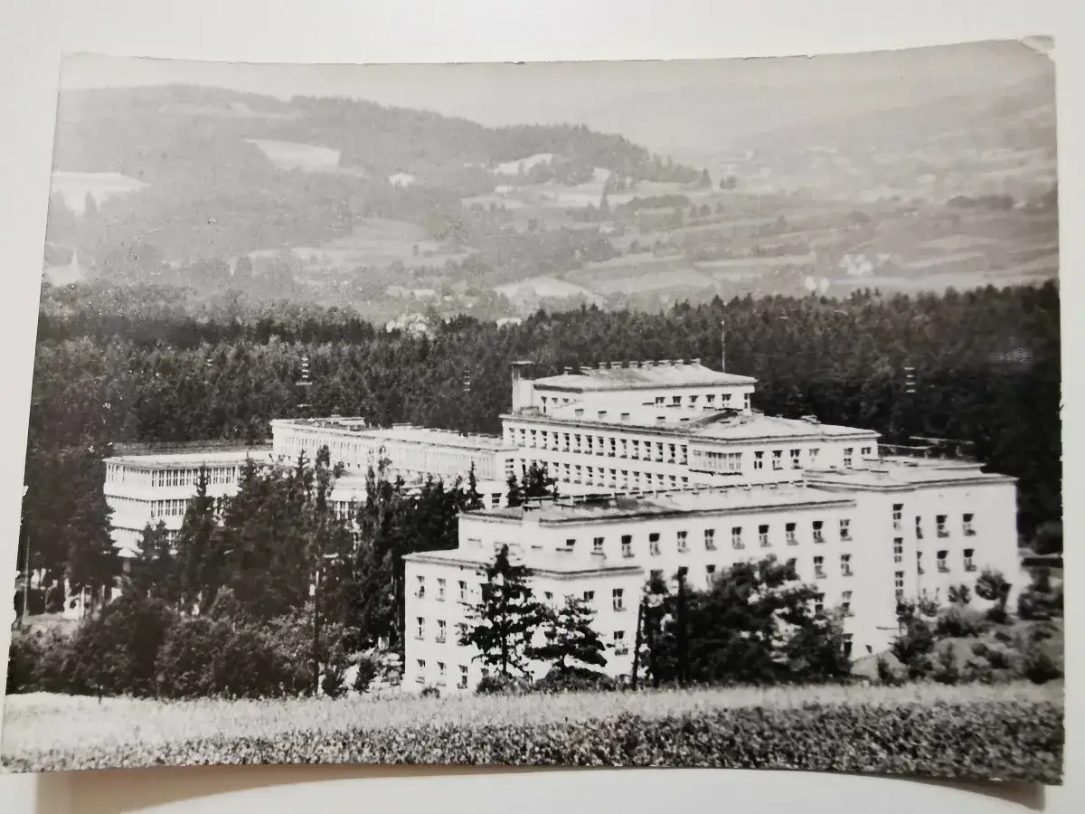 Duży budynek sanatorium w otoczeniu drzew, na tle pagórkowatego krajobrazu.