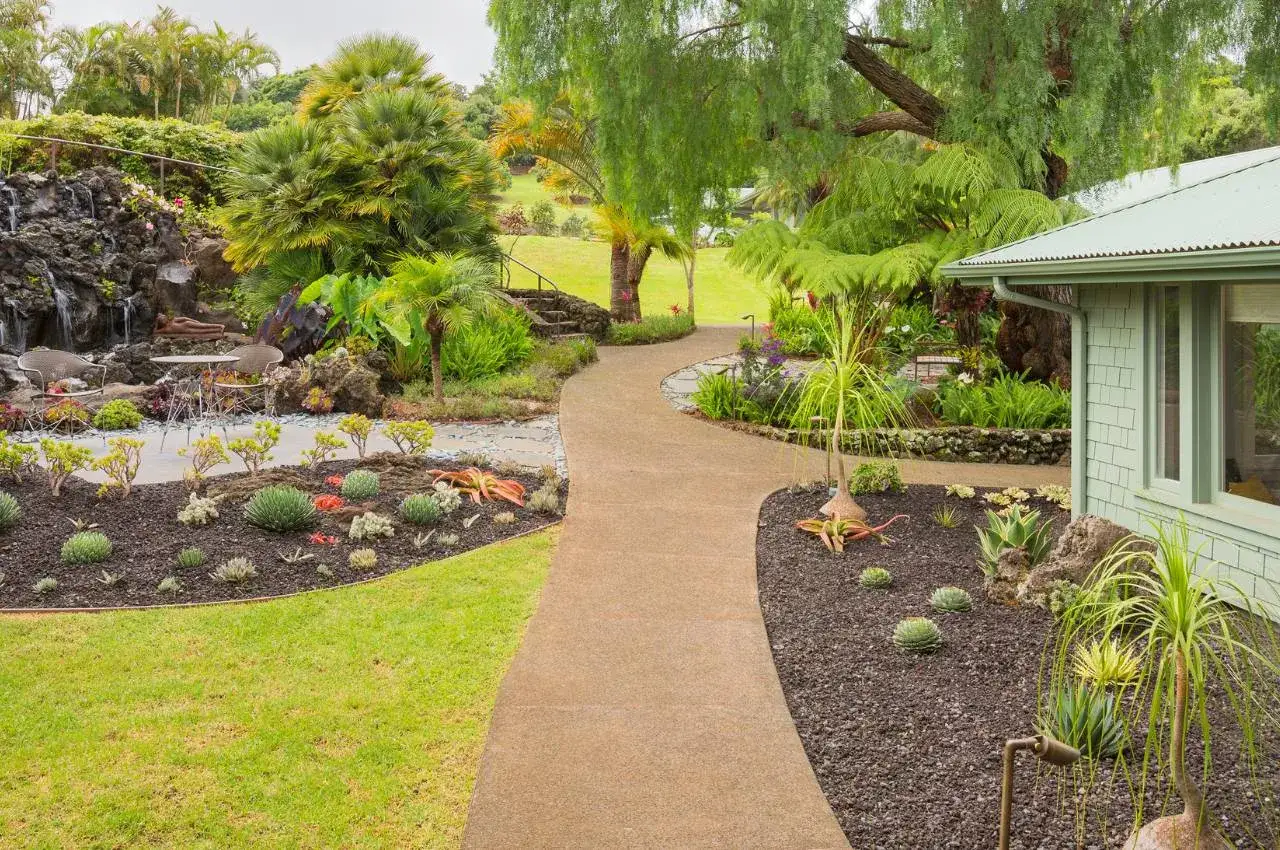 Piękna rabata żwirowa aranżacje z wodospadem, palmami i egzotyczną roślinnością. Ścieżka prowadzi do domu.