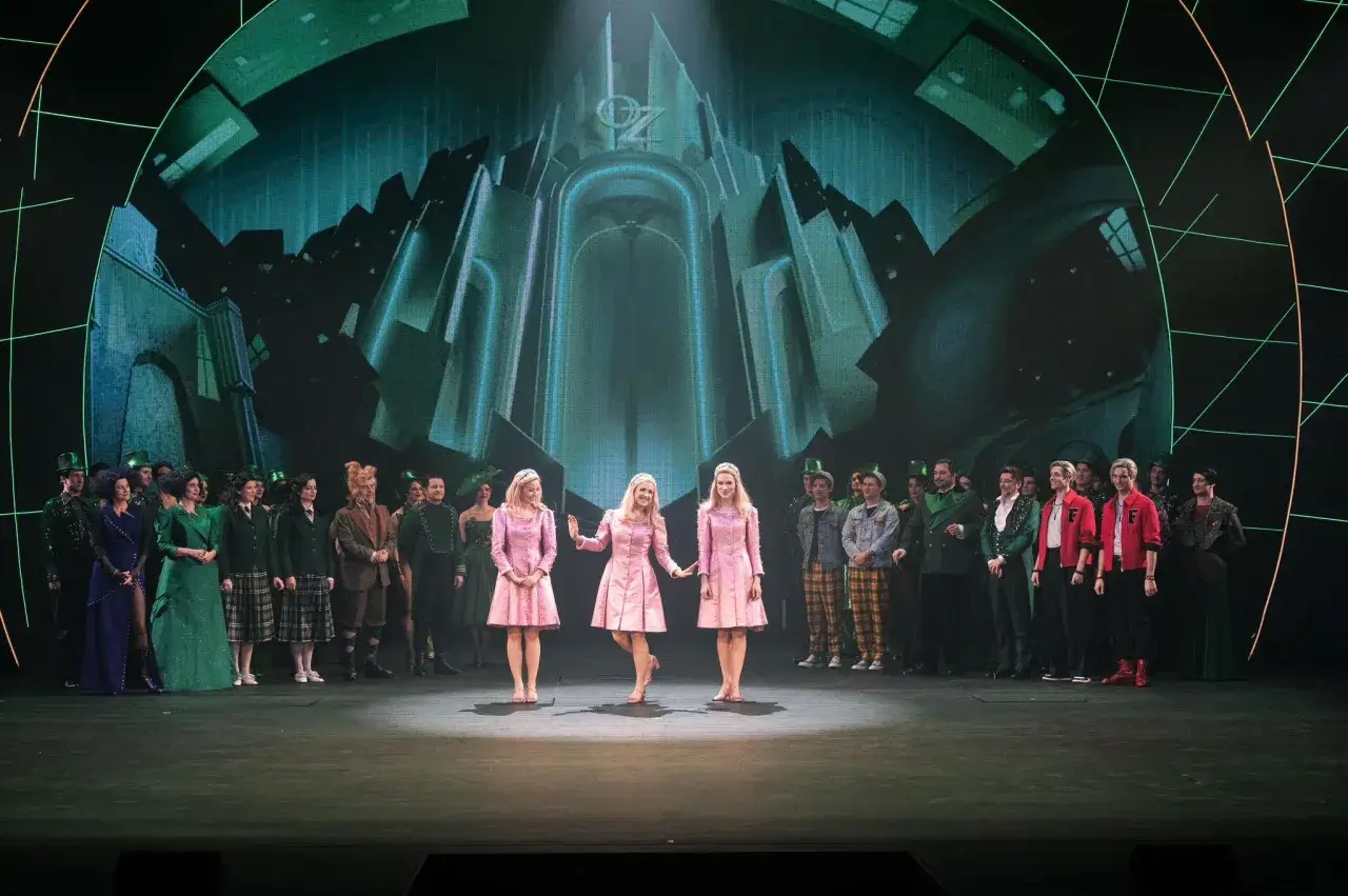 Trzy dziewczyny w różowych sukienkach na scenie, w tle zielony pałac Oz. To scena z musicalu "Wicked", który ma imponujący czas trwania.