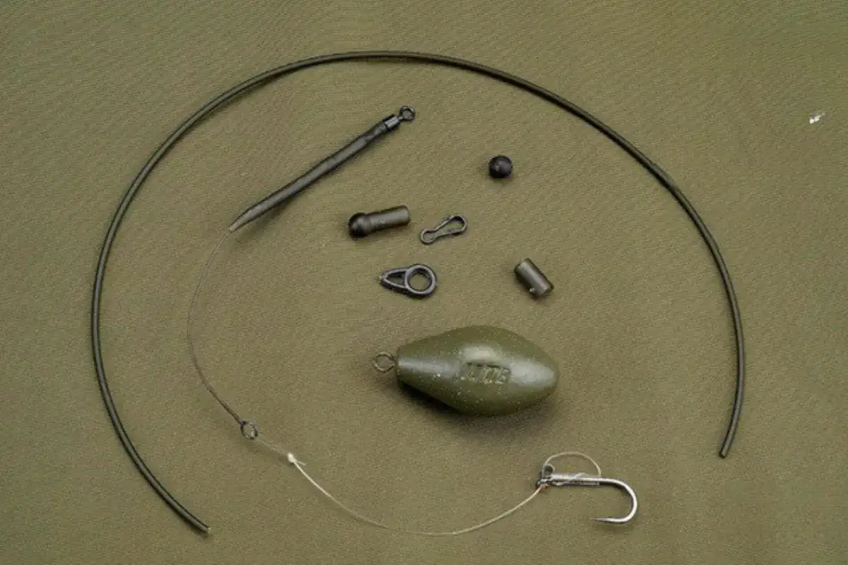 Akcesoria wędkarskie: ciężarek, haczyk, żyłka i elementy montażowe na zielonym tle.