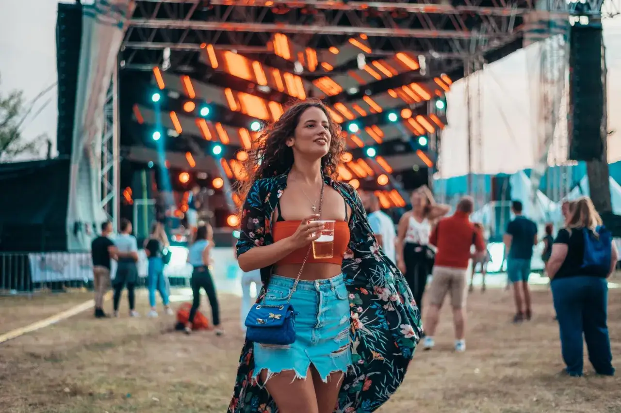 Dziewczyna w kwiecistej narzutce i jeansowej spódniczce, z drinkiem w ręku, pokazuje, jak ubrać się na koncert.