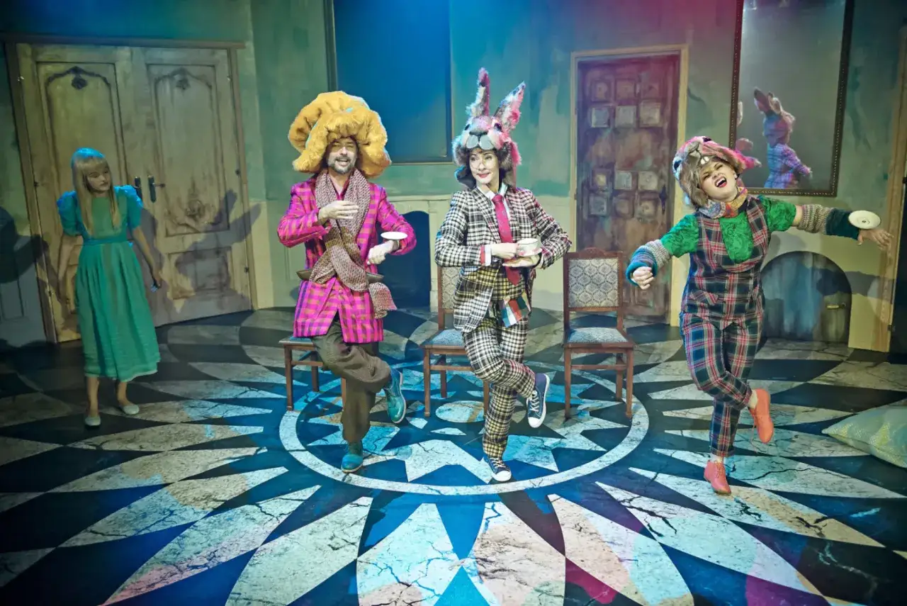 Aktorzy w strojach z "Alicji w Krainie Czarów" tańczą na scenie. W tle drzwi i lustro.