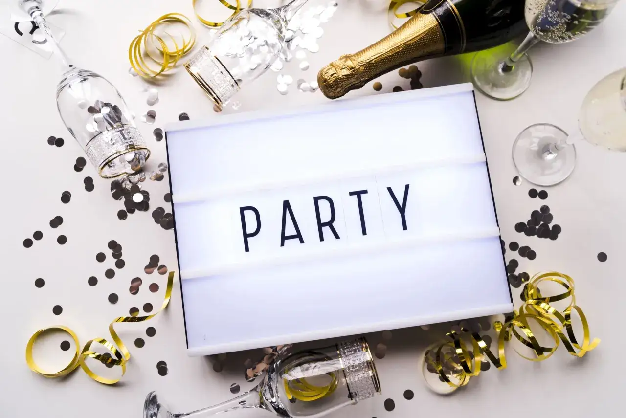 Świętowanie zaczyna się od zaproszeń! Na stole leżą kieliszki, szampan i konfetti, a tablica świetlna głosi "PARTY".