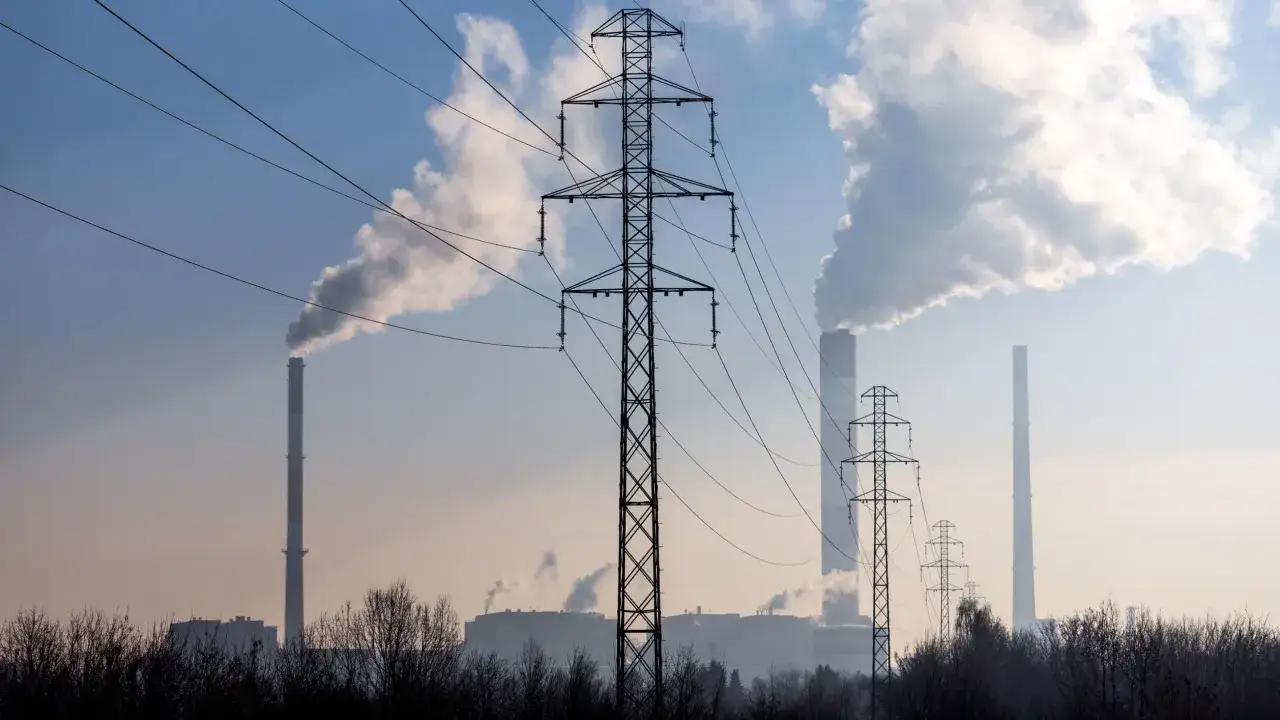 Linie energetyczne i dymiące kominy elektrowni na tle błękitnego nieba. W tle widać kilka wież transmisyjnych i budynki przemysłowe.