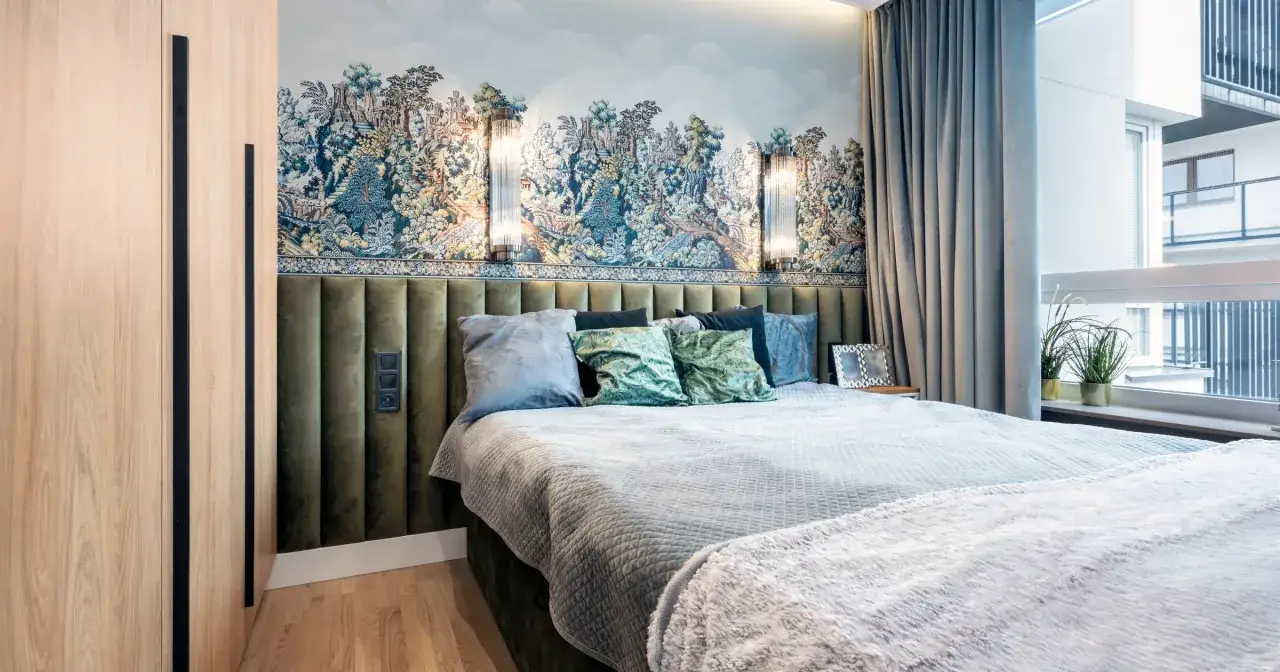 Tapicerowane panele na ścianie nad łóżkiem, przyklejone na tapetę z motywem krajobrazu, tworzą przytulną sypialnię.