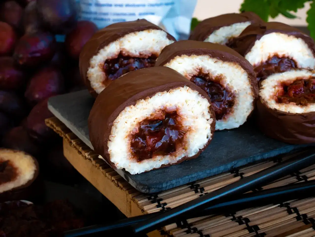 Słodkie rolki sushi na słodko, oblane czekoladą, z owocowym nadzieniem. Obok ciemne owoce i pałeczki.