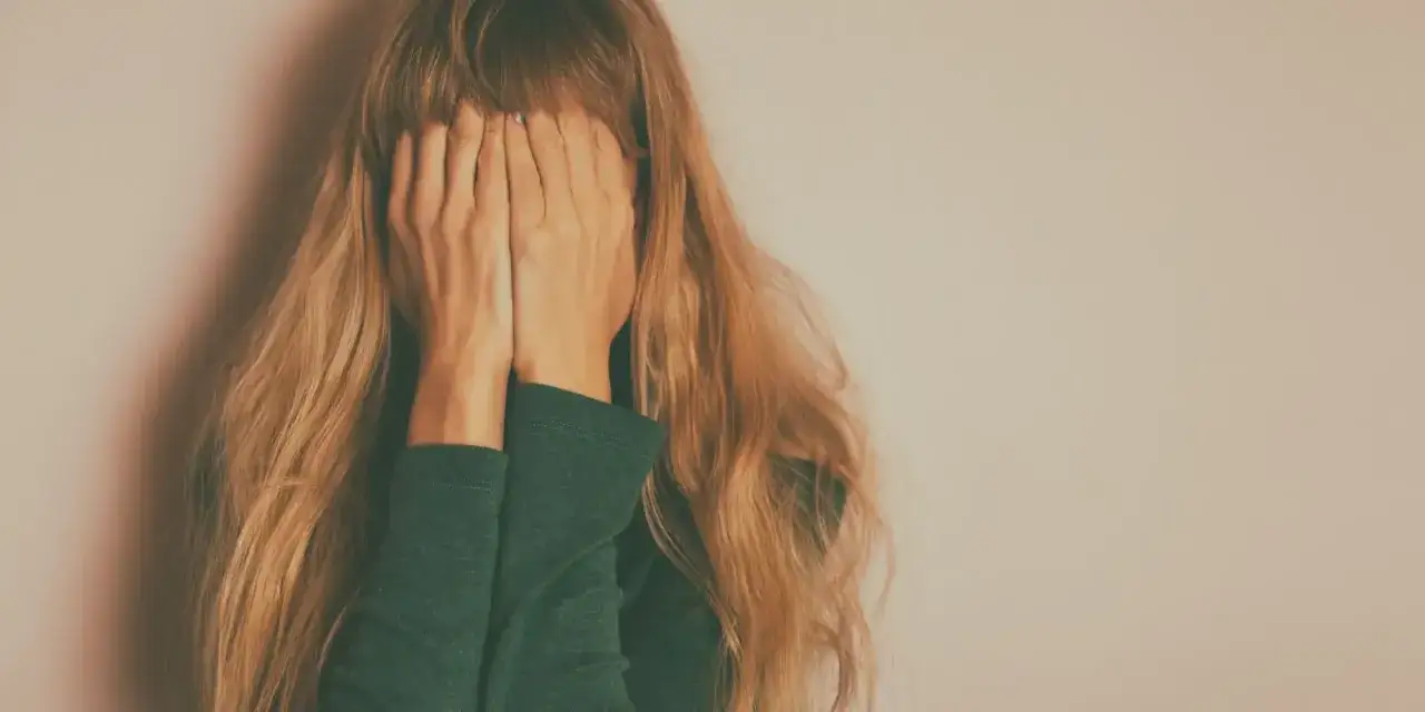 Kobieta z długimi blond włosami zakrywa twarz dłońmi na tle jasnej ściany.