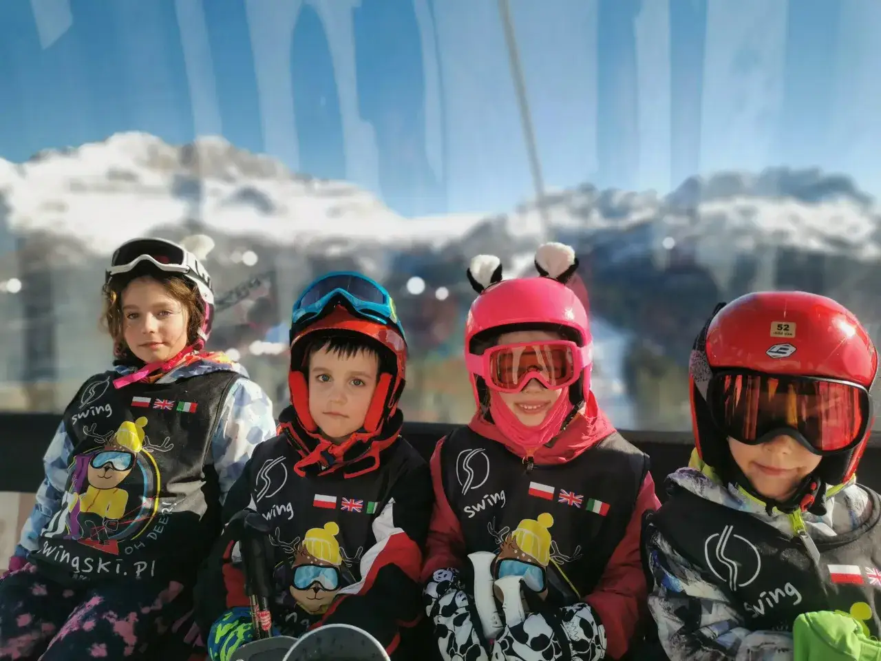 Cztery dzieci w strojach narciarskich i goglach siedzą w kolejce linowej, patrząc w stronę obiektywu.