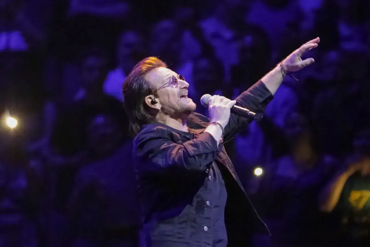 Bono z U2 śpiewa na koncercie, zapowiadając u2 nowa płyta. Tłum w ciemnościach reaguje na jego występ.