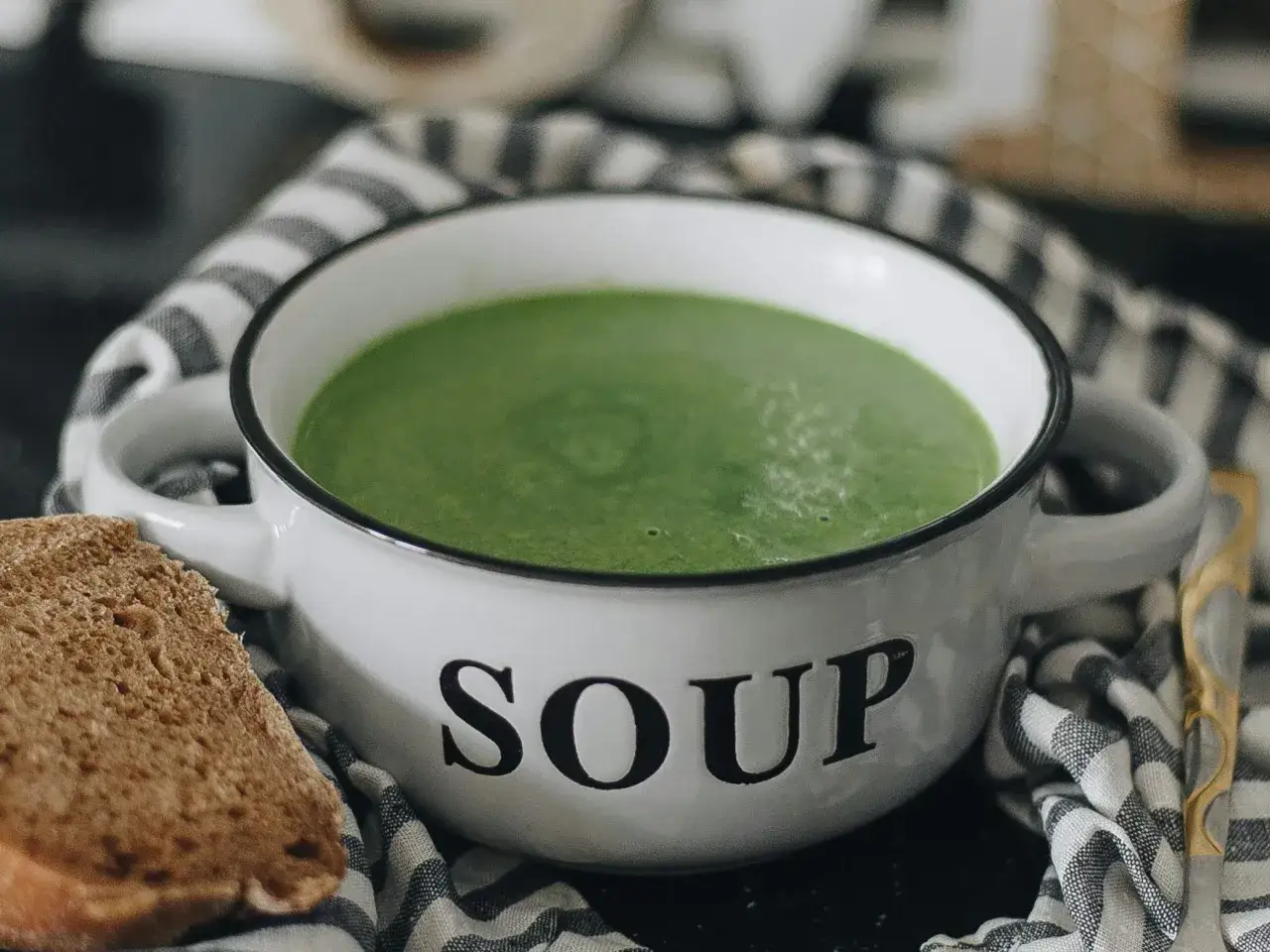 Zielony kremowy zupa w białej misce z napisem "SOUP", obok kromka chleba. Idealne rozwiązanie, jak mrozić zupy na później.