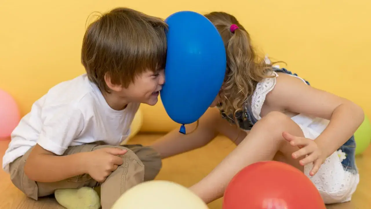 Dwoje dzieci z baloniem między twarzami, otoczeni kolorowymi balonami.