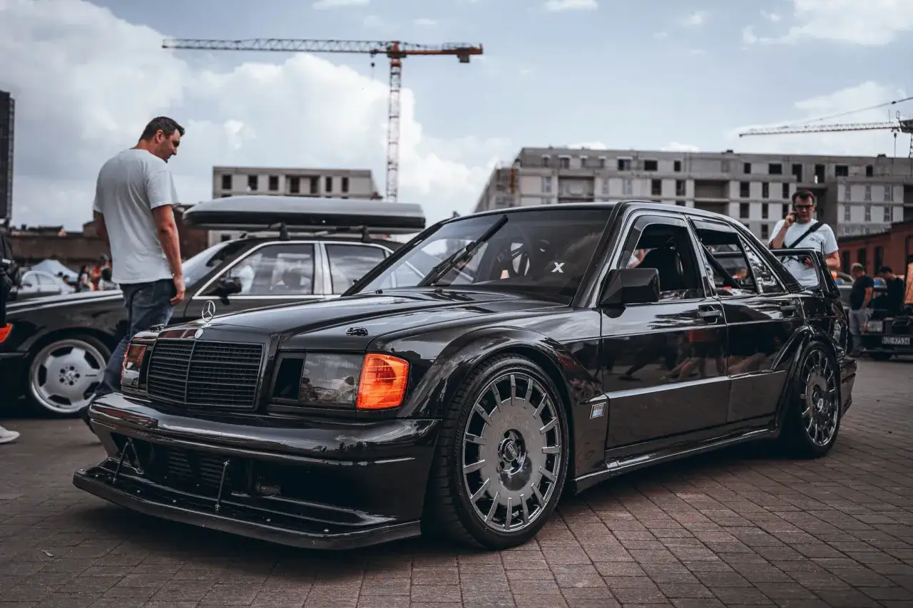Czarny Mercedes 190 z agresywnym body kitem Evo, lśniącym na zlotowym placu.