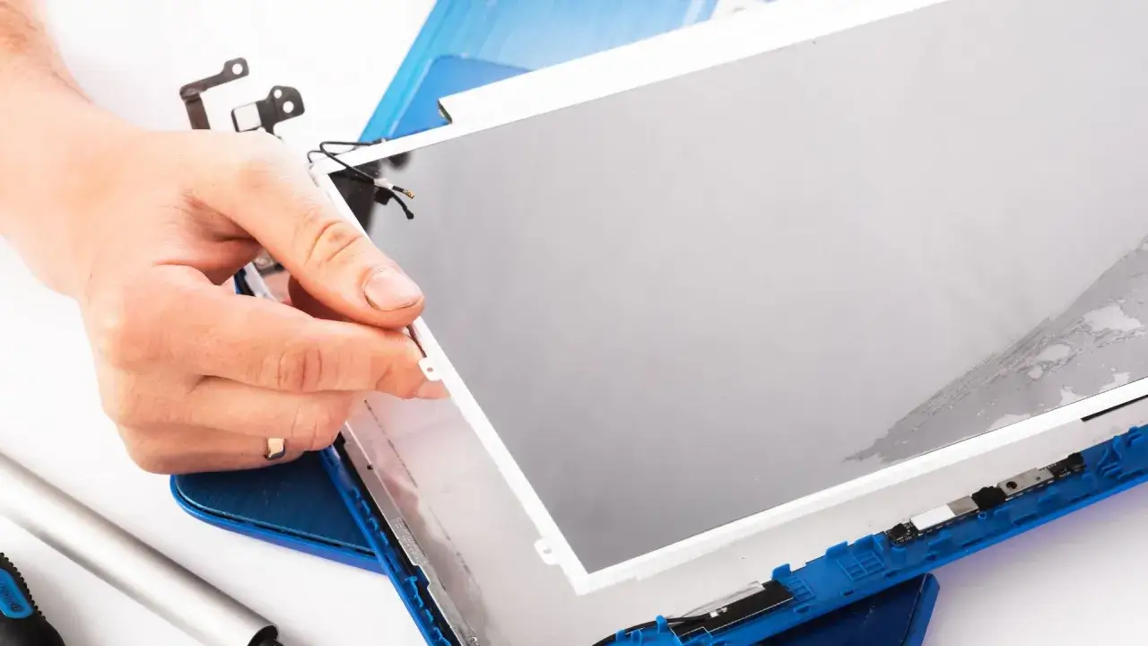 Dłoń trzyma matrycę w laptopie podczas naprawy. Widać narzędzia i niebieską obudowę.