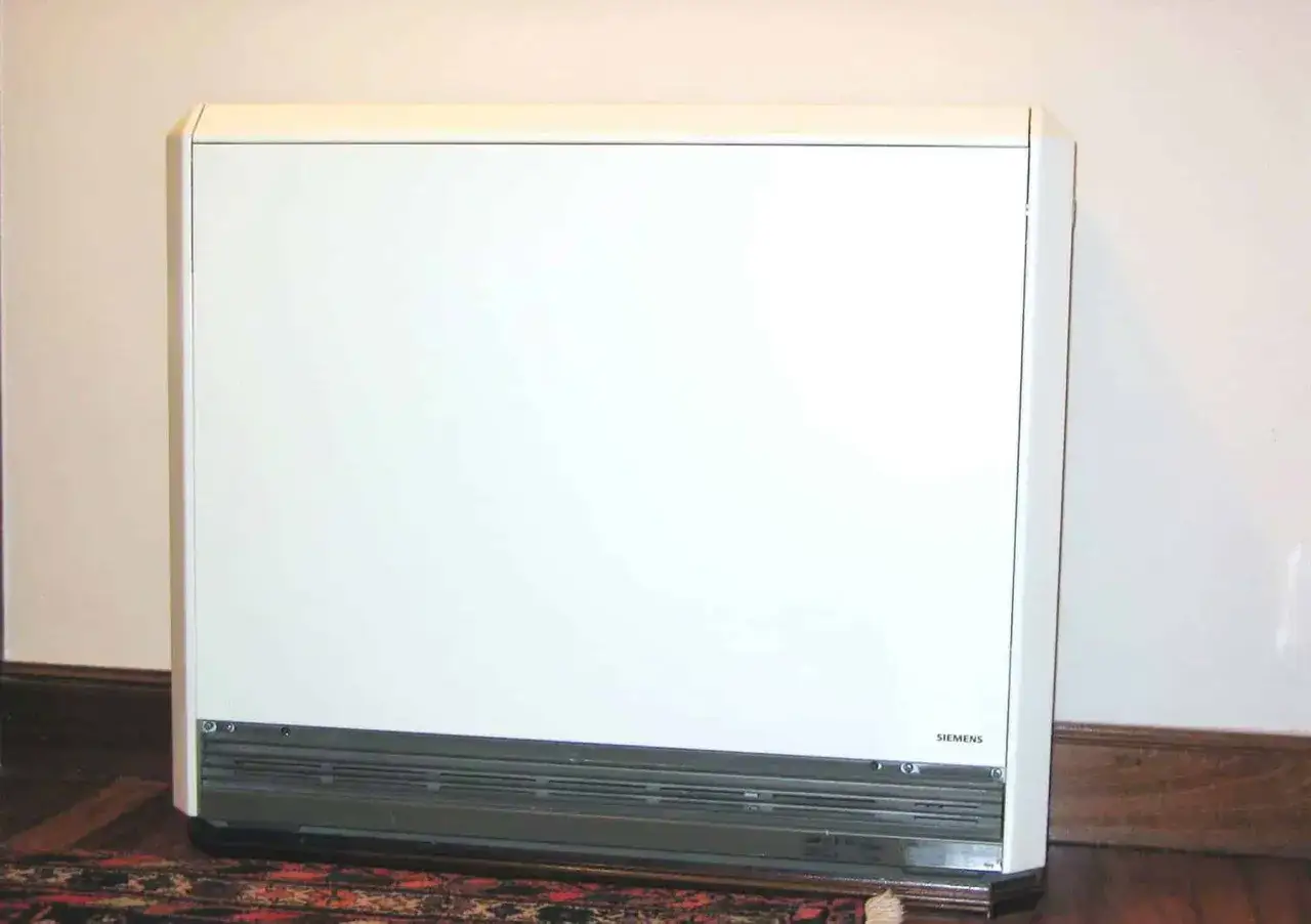 Biały, prostokątny piec akumulacyjny elektryczny marki Siemens stoi na drewnianej podłodze, obok dywanu.