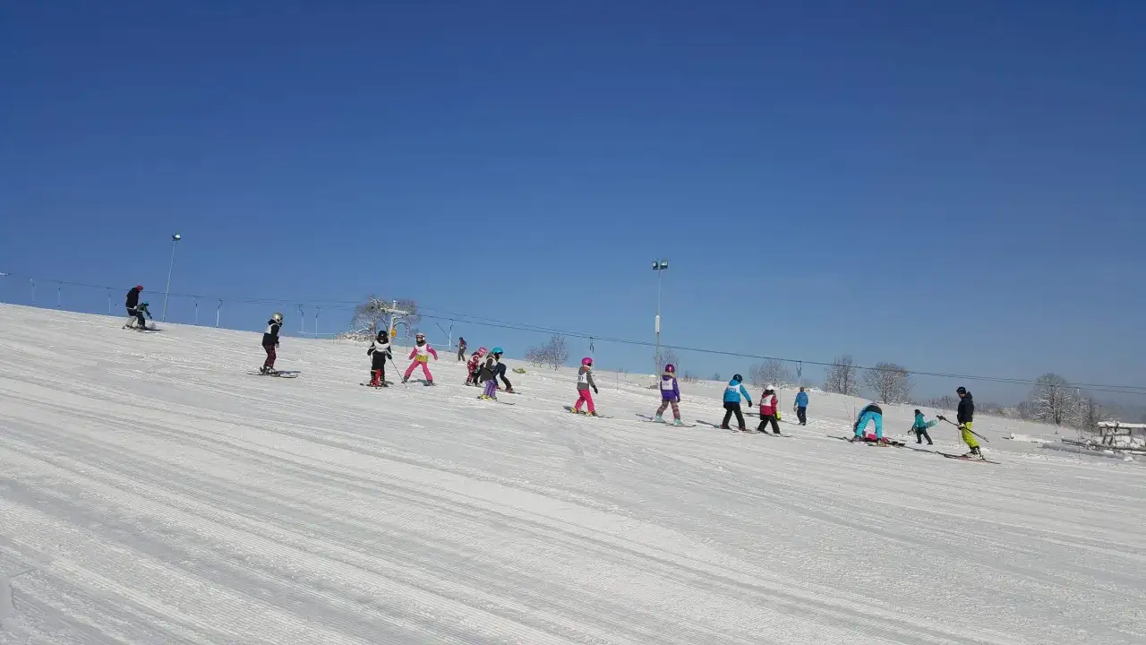 Dzieci uczą się jeździć na nartach na stoku Budzowski Wierch. Słońce świeci, a niebo jest błękitne.