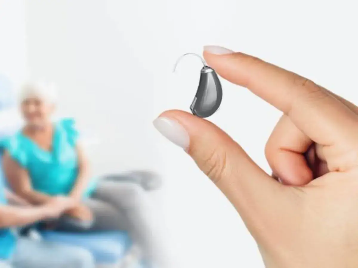 Dłoń trzyma aparat słuchowy. W tle starsza kobieta.