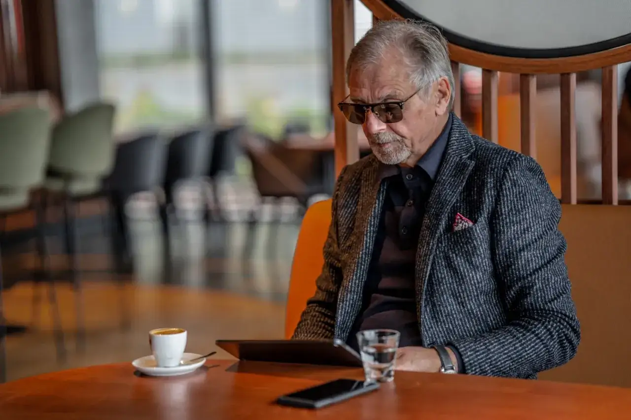 Elegancki starszy pan w okularach, z kawą i tabletem, zastanawia się, co kupić starszej osobie.