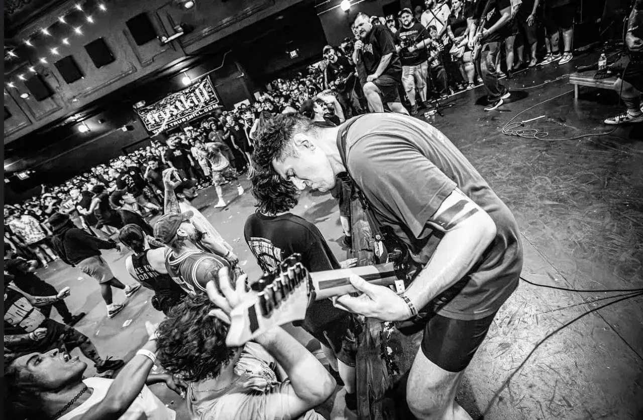 Energetyczny koncert w klubie. Czarno białe zdj. z tłumem szalejącym pod sceną, gdzie gitarzysta pochyla się nad instrumentem.