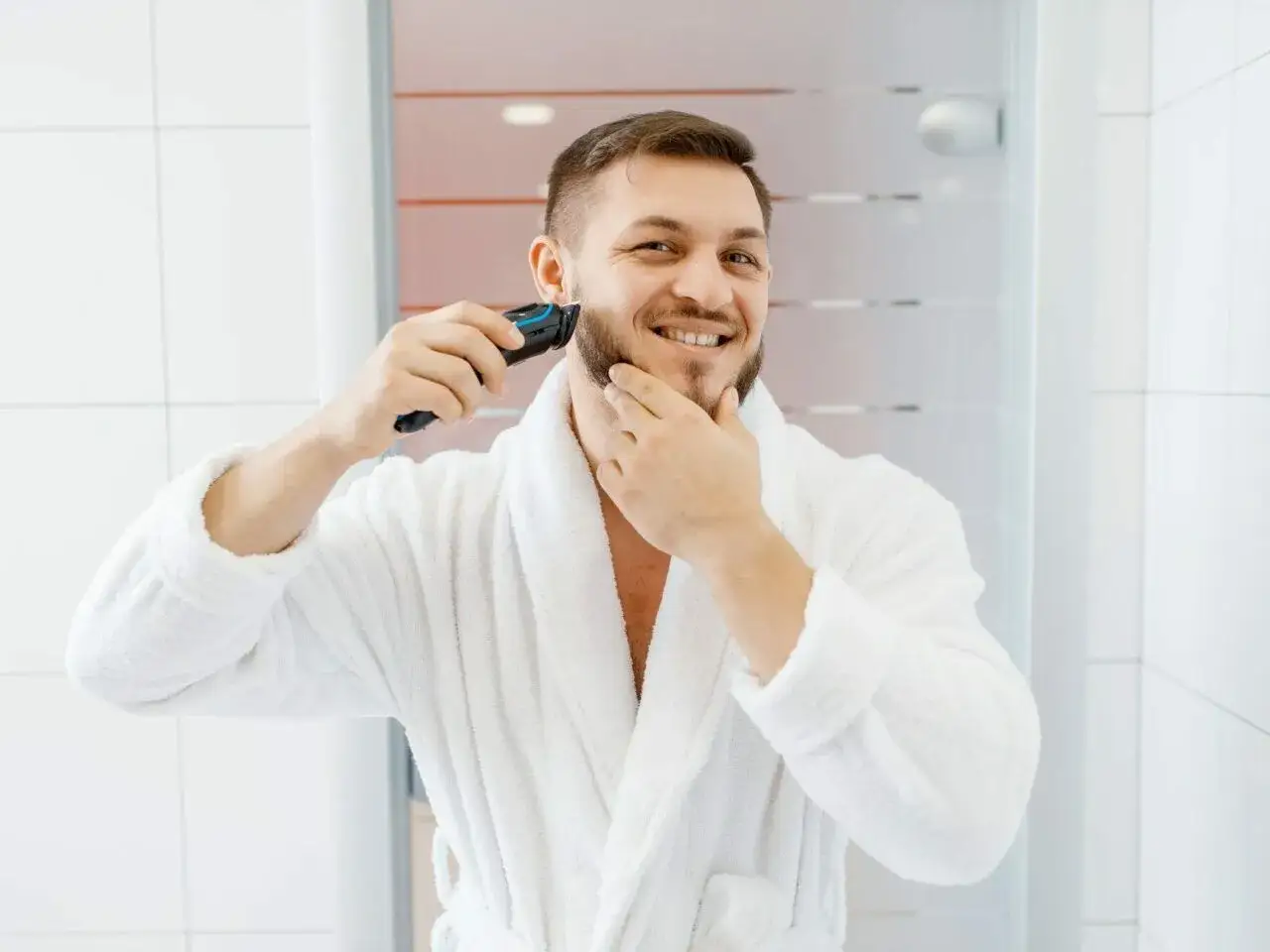 Uśmiechnięty mężczyzna w białym szlafroku goli brodę trymerem w łazience.