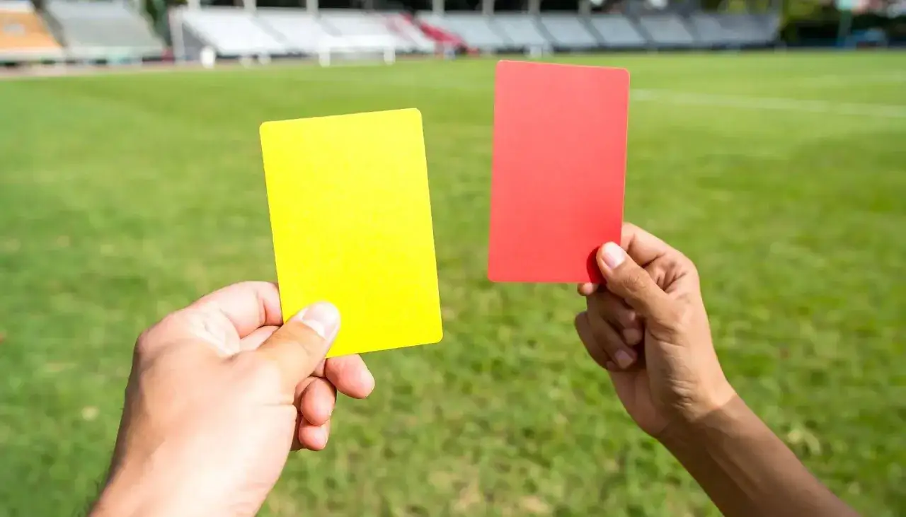 Sędzia pokazuje żółtą kartkę, która oznacza ostrzeżenie w piłce nożnej. Obok czerwona kartka.