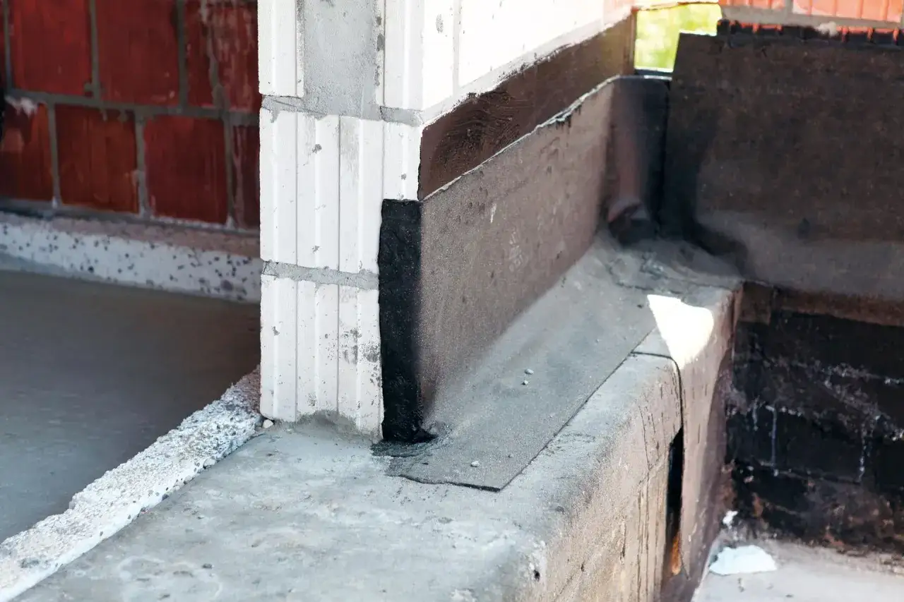 Fragment budowy z widoczną izolacją przeciwwilgociową, czyli papą na fundament. Widać beton, pustaki i czarną masę izolacyjną.