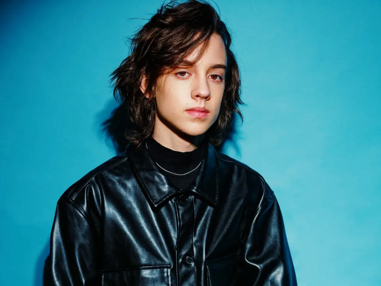 Młody mężczyzna w czarnej skórzanej kurtce, z burzą brązowych włosów, patrzy wprost, jakby czekał na znak, kto śpiewa.