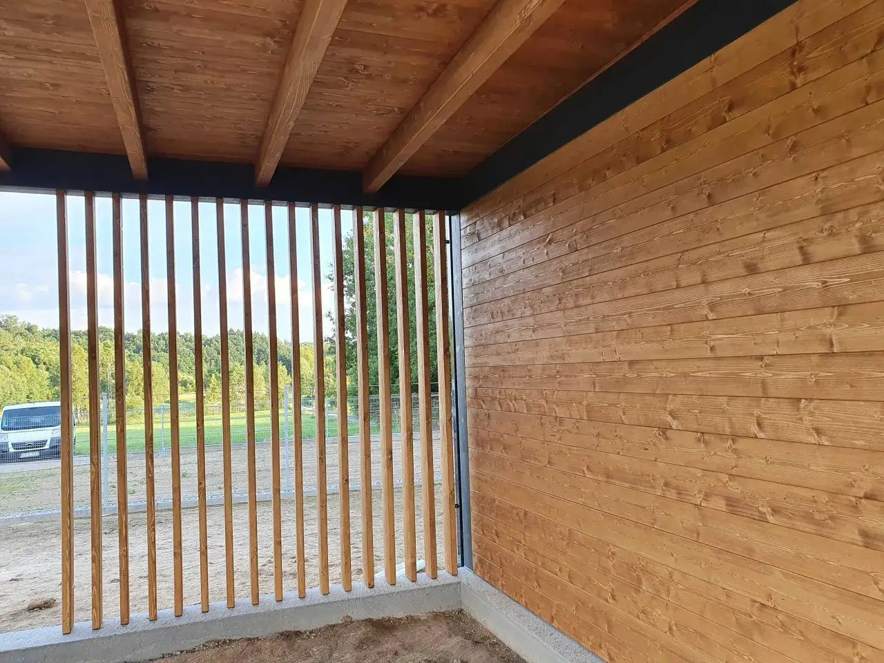 Drewniane deski na ścianach altany tworzą ciepły klimat. Pionowe listwy zapewniają widok na zieleń i niebo.