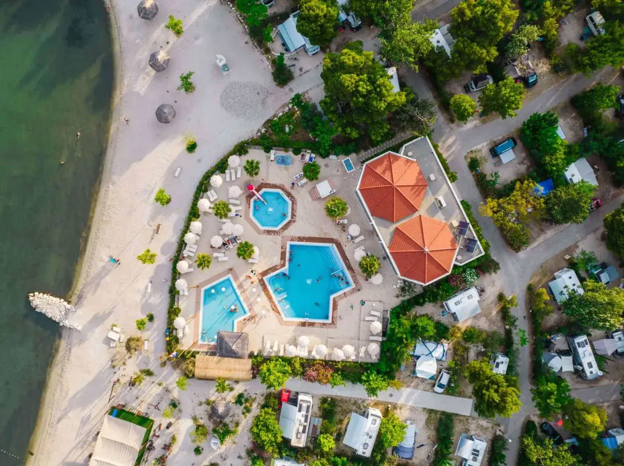 Widok z lotu ptaka na kemping z basenami i plażą. Widać tu podział na strefy: plażową, basenową i kempingową z kamperami.