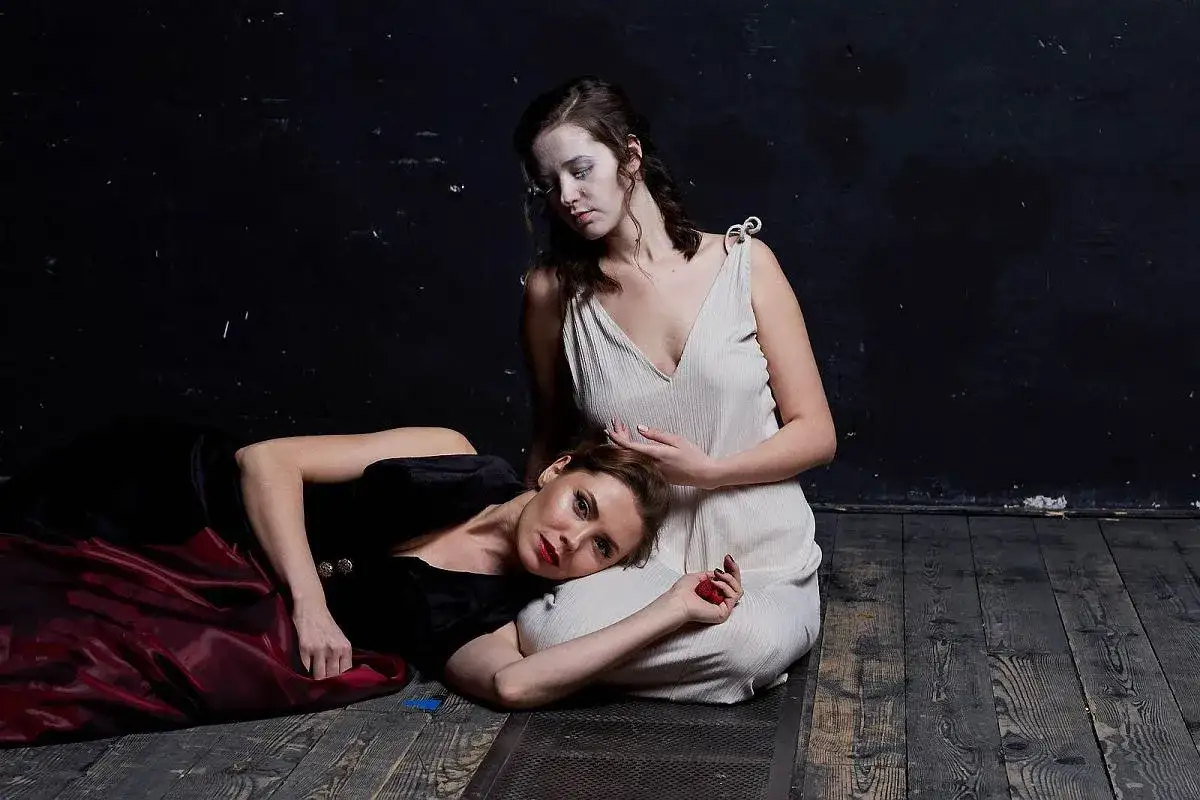 Dwie kobiety w sukniach, jedna leży, druga siedzi, na drewnianej podłodze.