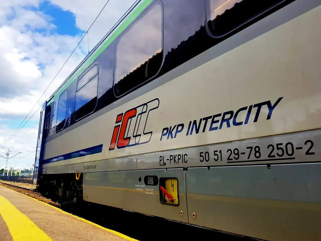 Pociąg PKP Intercity z napisem "Warszawa Poznań" na tle błękitnego nieba z chmurami.