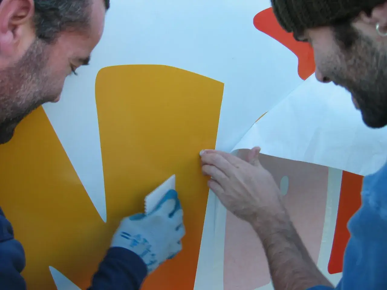 Dwóch mężczyzn pokazuje, jak nakleić naklejkę na ścianę. Jeden wygładza ją raklem, drugi odkleja papier.