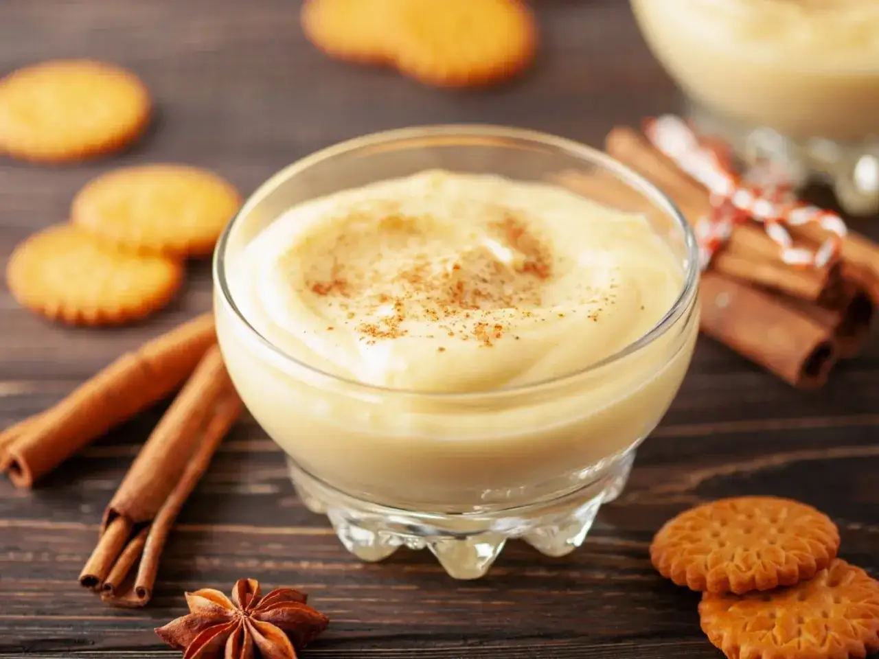 Kremowy pudding w szklanej miseczce posypany cynamonem, obok ciasteczka i laski cynamonu.