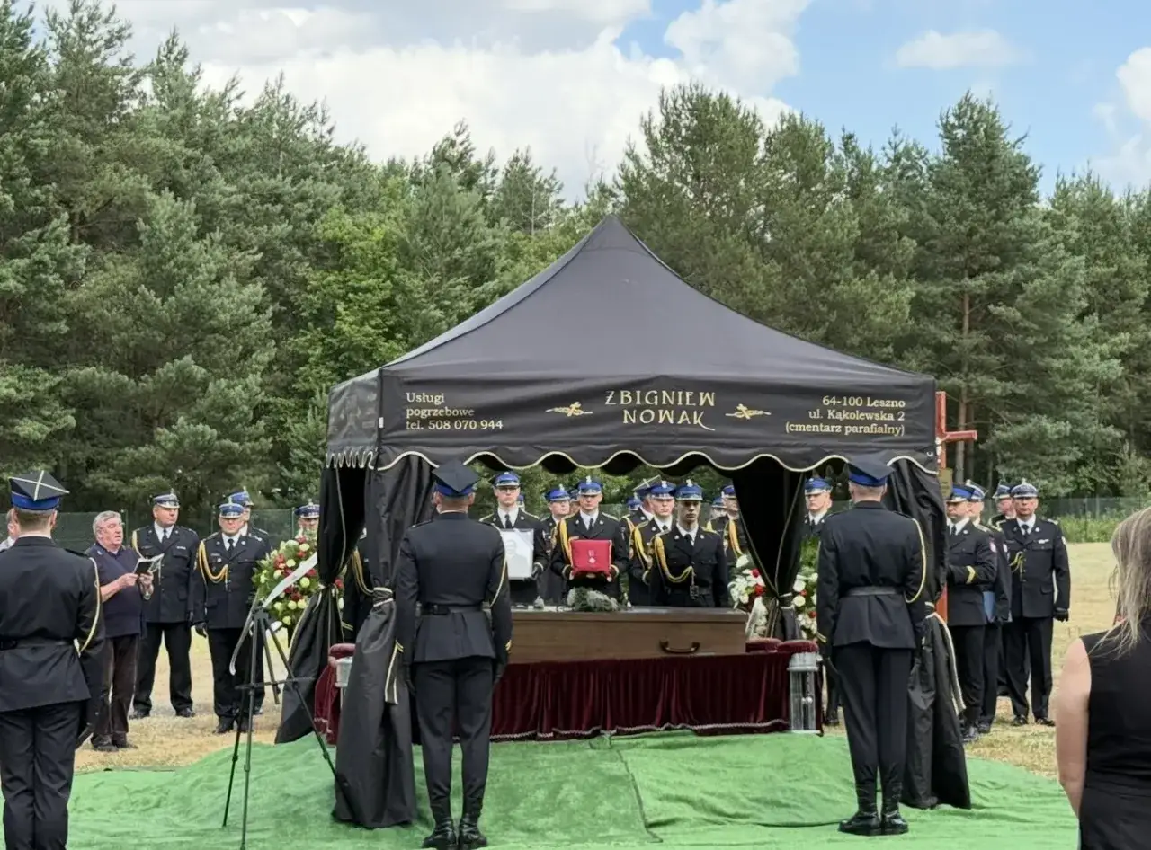 Uroczystość pogrzebowa pod namiotem z trumną, w otoczeniu osób w mundurach.