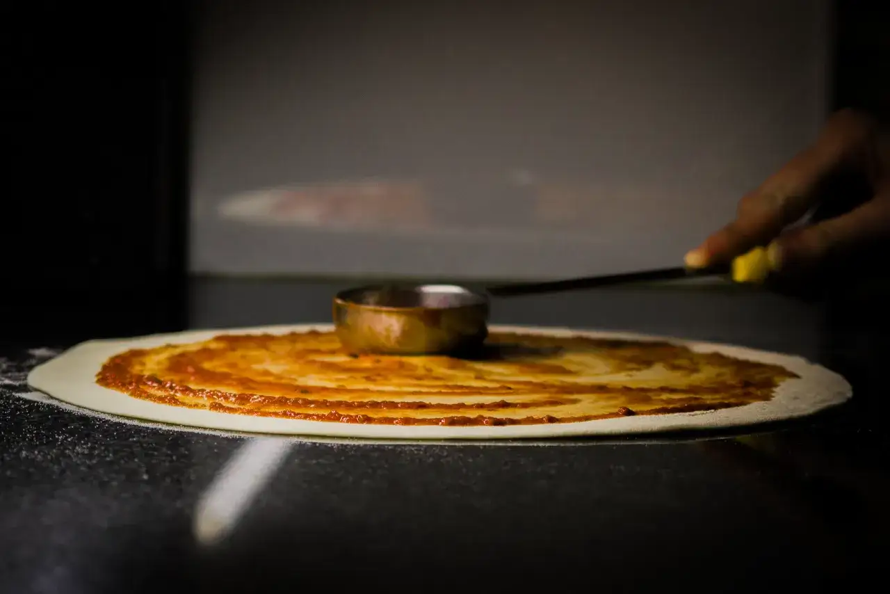Ręka rozprowadza sos pomidorowy na cieście, tworząc idealny zaczyn na pizzę.