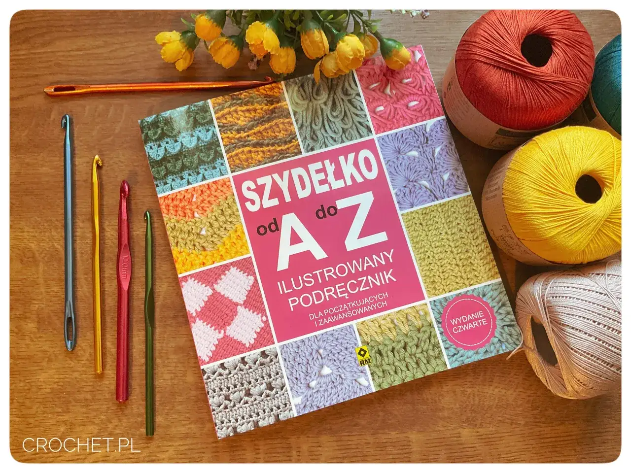 Książka "Szydełko od A do Z" z kolorowymi motywami, obok niej kilka szydełek, w tym jedno długie, oraz kłębki włóczki.