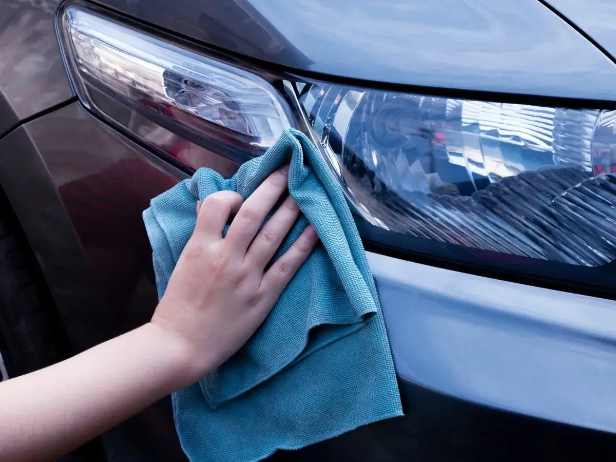 Dłoń z niebieską mikrofibrą wyciera reflektor ciemnego samochodu.