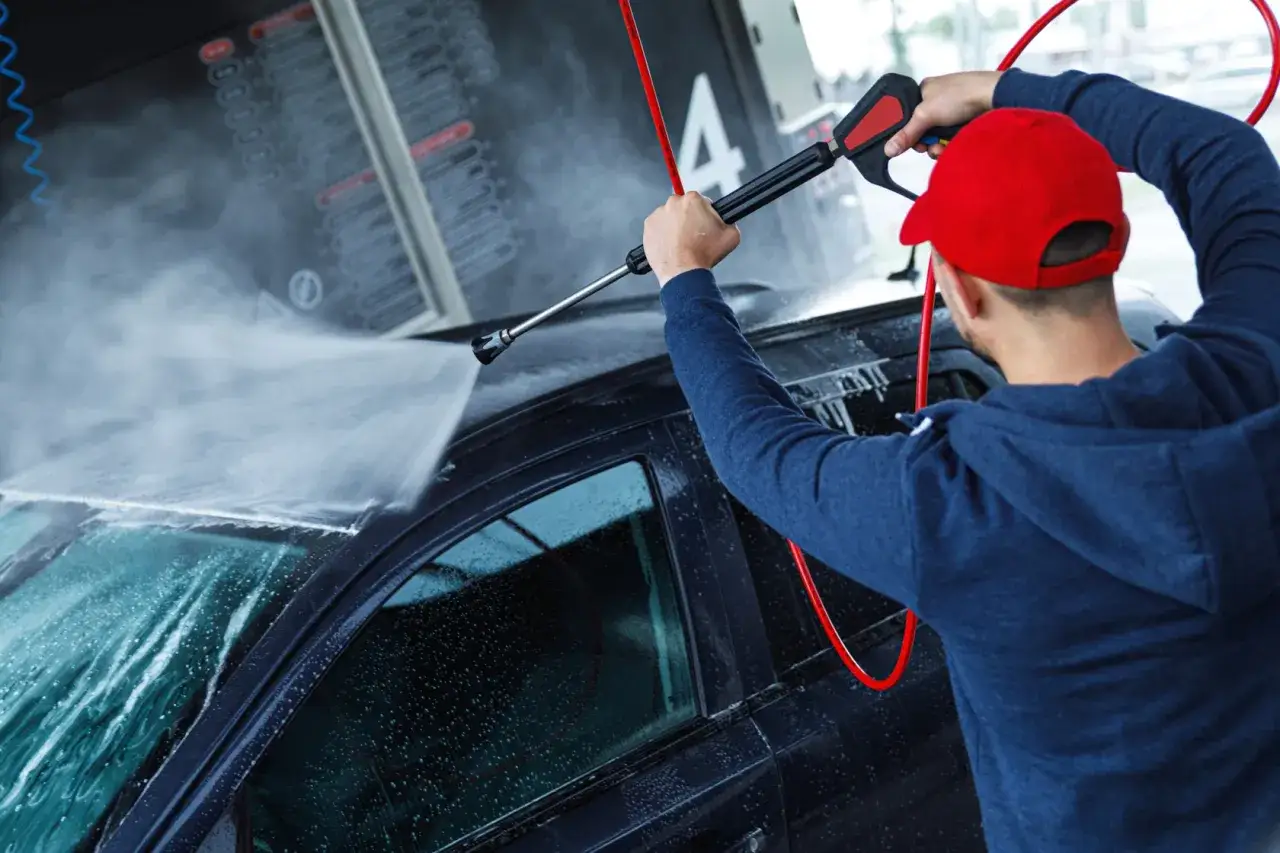Mężczyzna w czerwonej czapce pokazuje, jak myć auto na myjni ręcznej, spryskując je wodą z myjki ciśnieniowej.