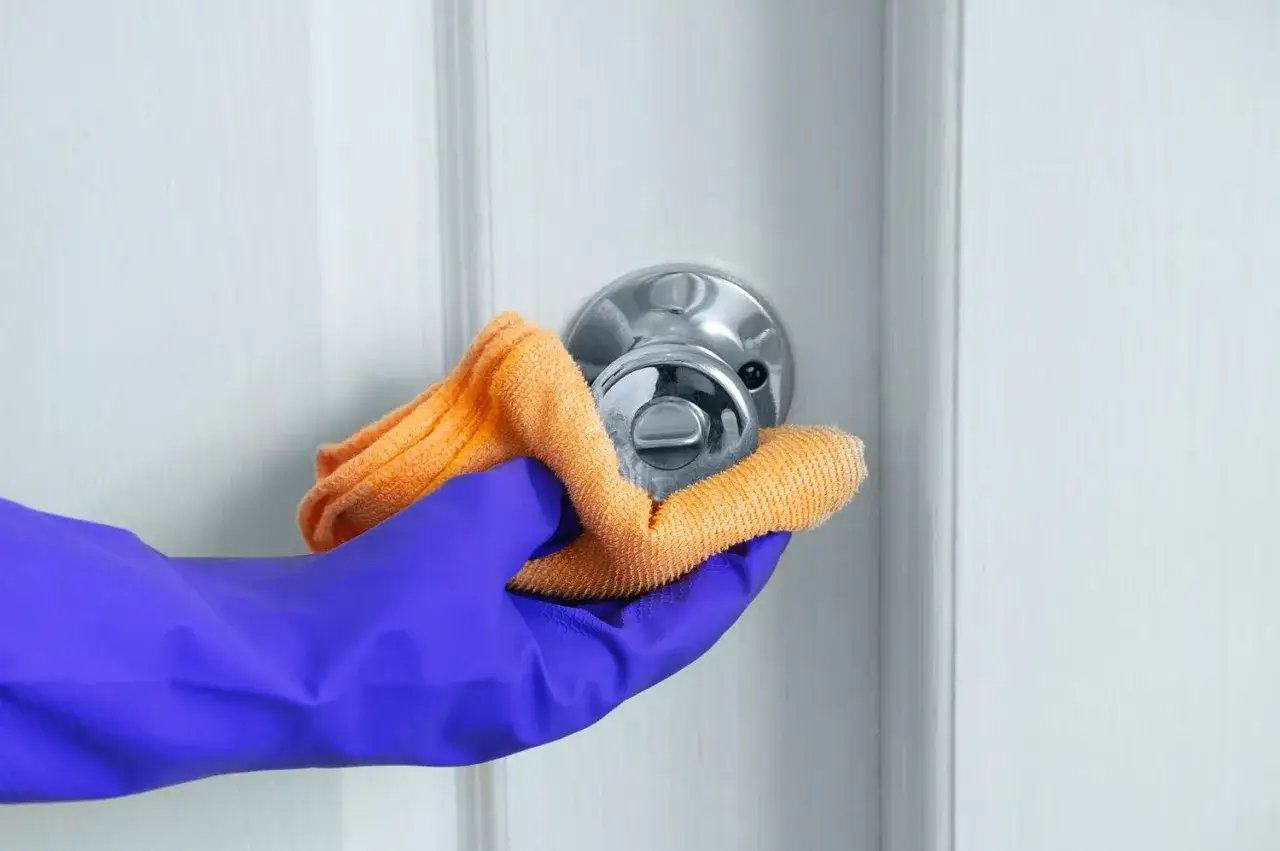 Ręka w fioletowej rękawiczce czyści klamkę pomarańczową ściereczką.